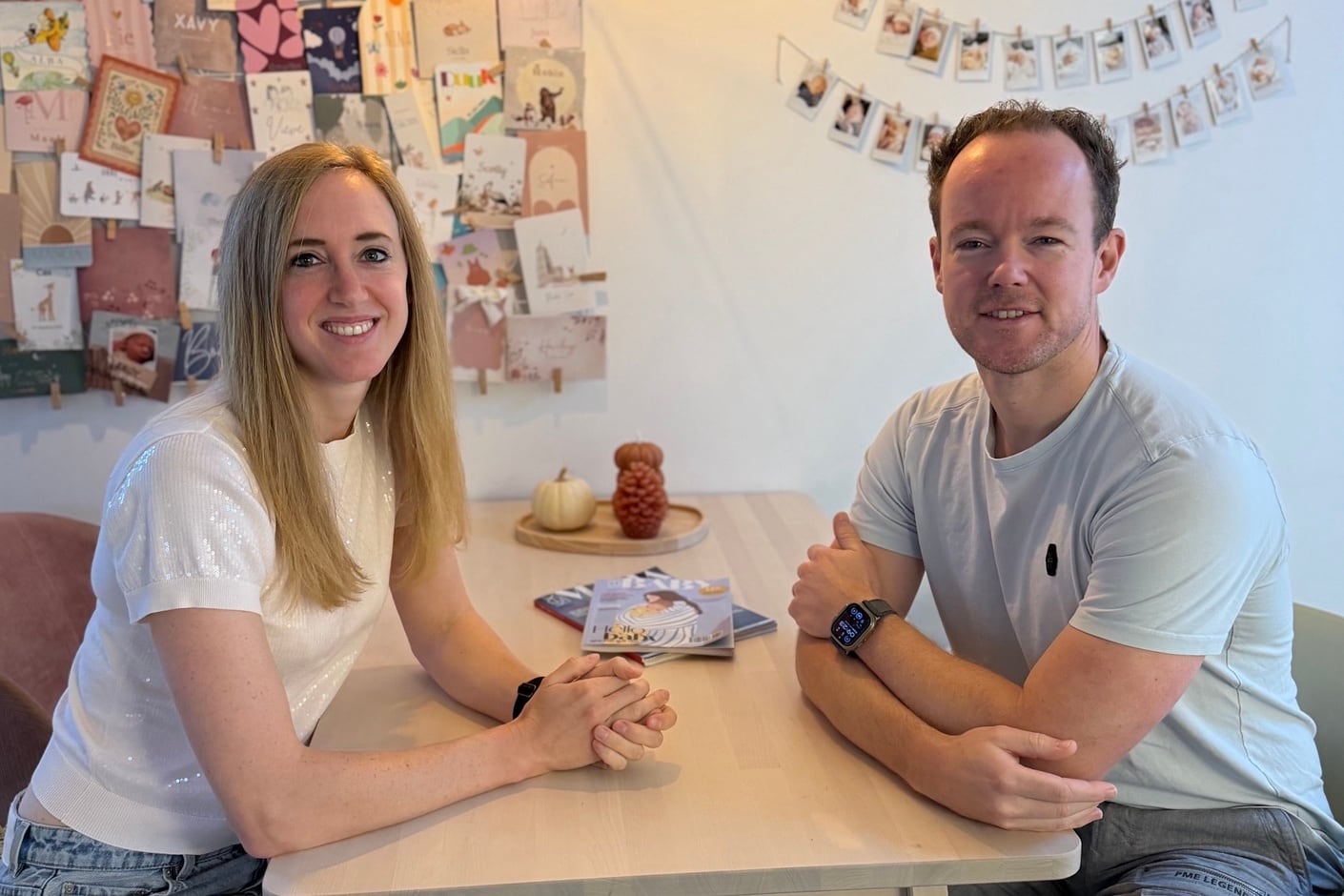 Jessica en Bram aan tafel, met geboortekaartjes en babyfoto’s aan de wand achter hen.